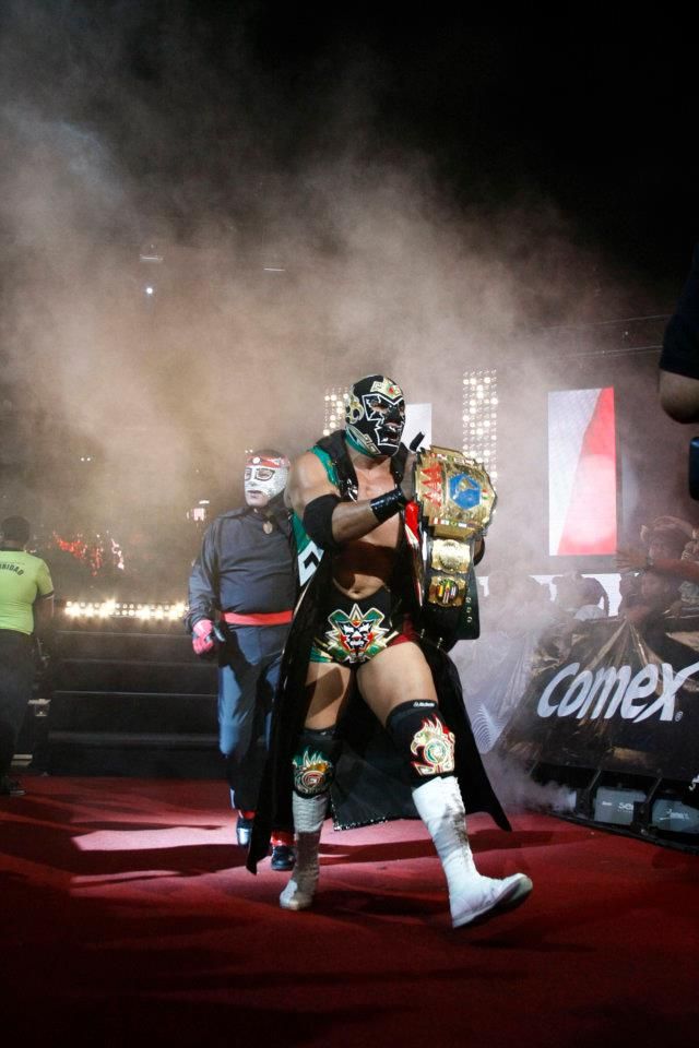 The image shows a man in a wrestling ring wearing a mask and holding a trophy, surrounded by a...