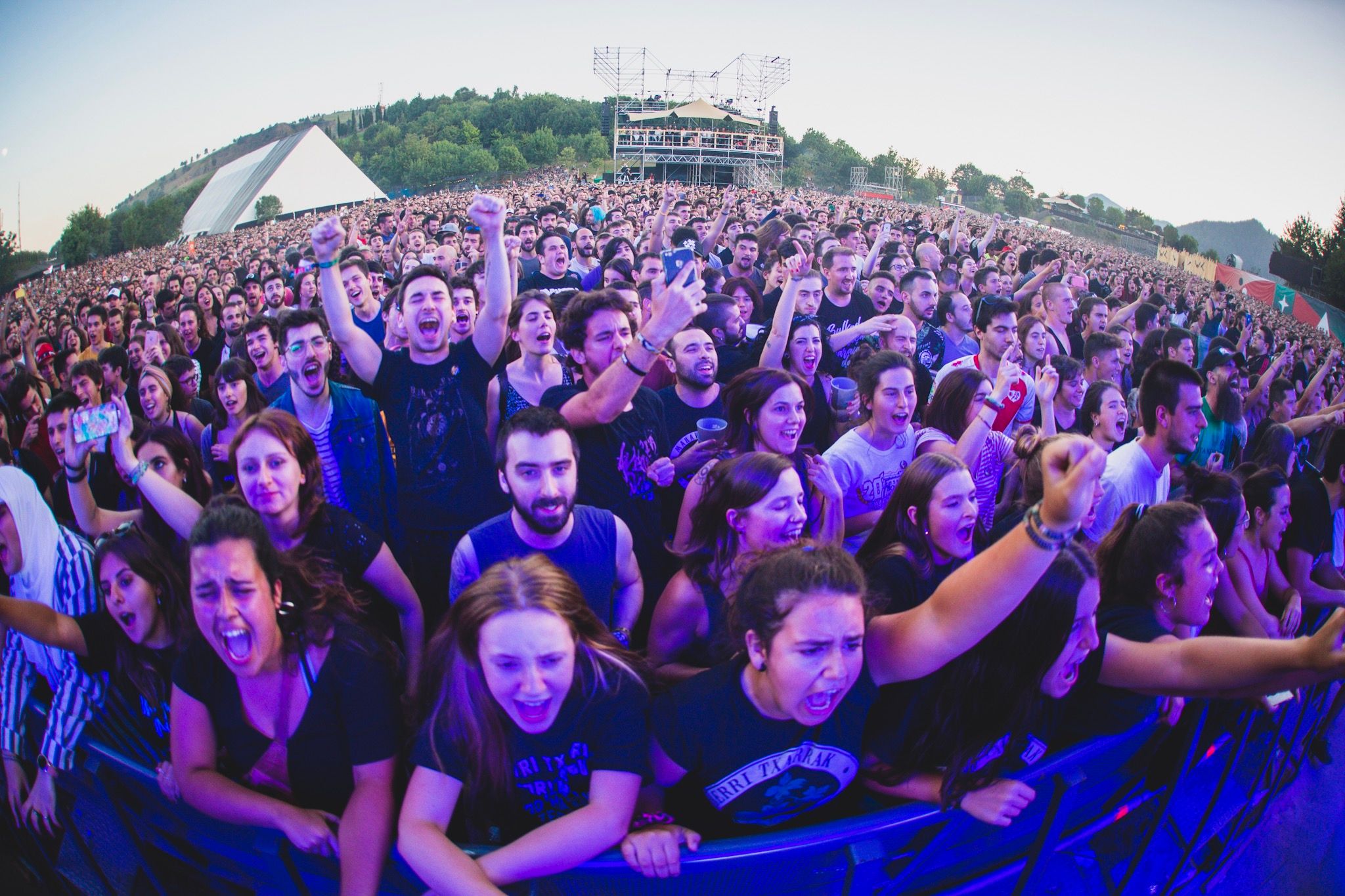 The image shows a large crowd of people at a music festival with their hands in the air, standing...