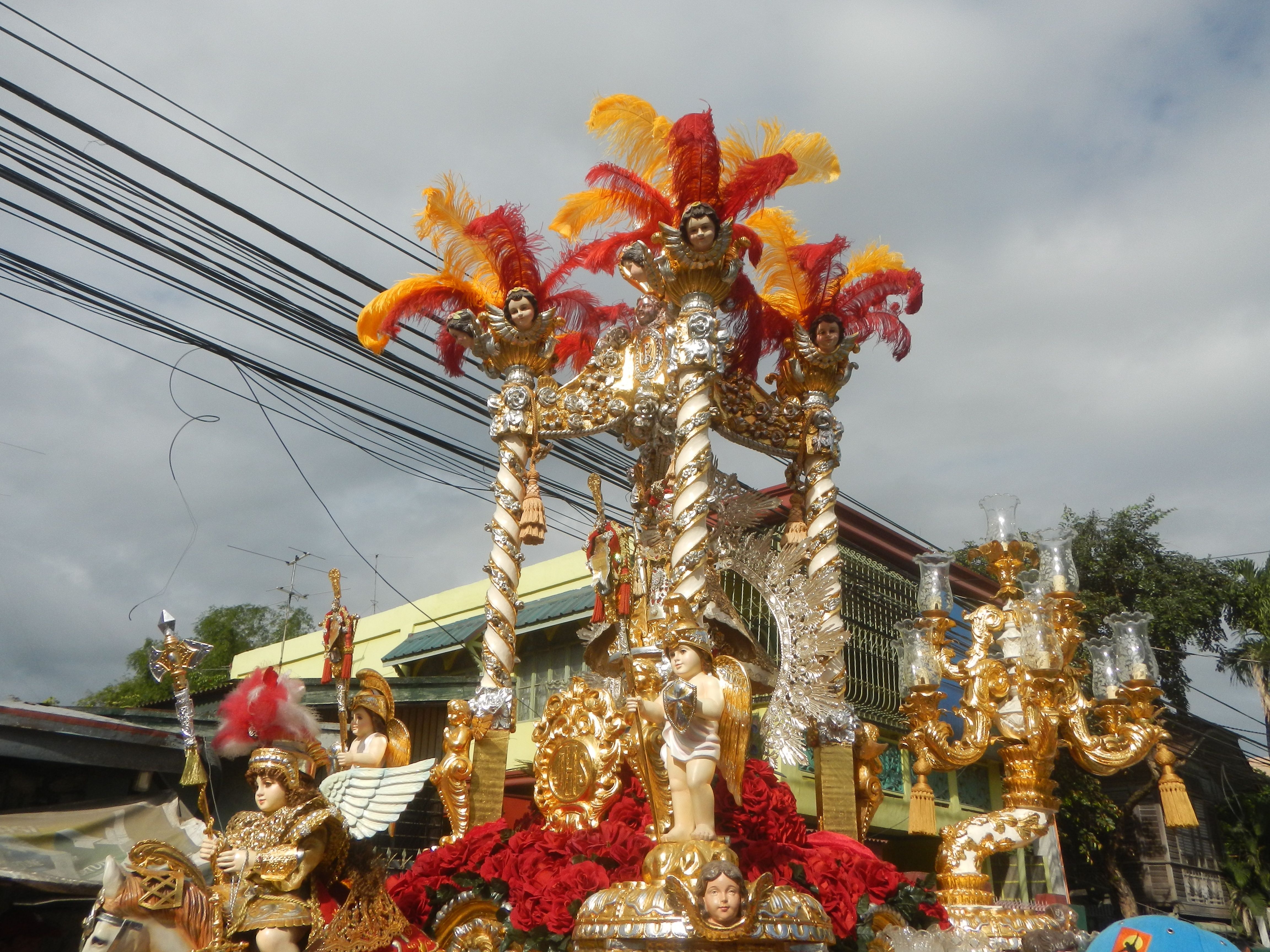 The image shows a float in a carnival parade with people on it, surrounded by buildings, trees,...