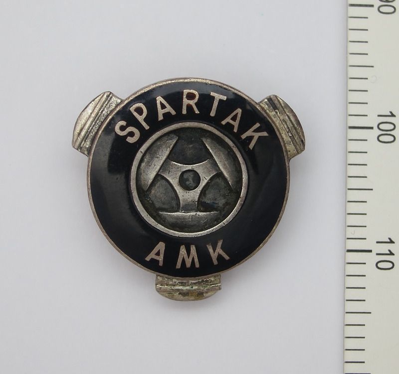 The image shows a spartak amk lapel pin on a white surface with a scale beside it. The pin is black...