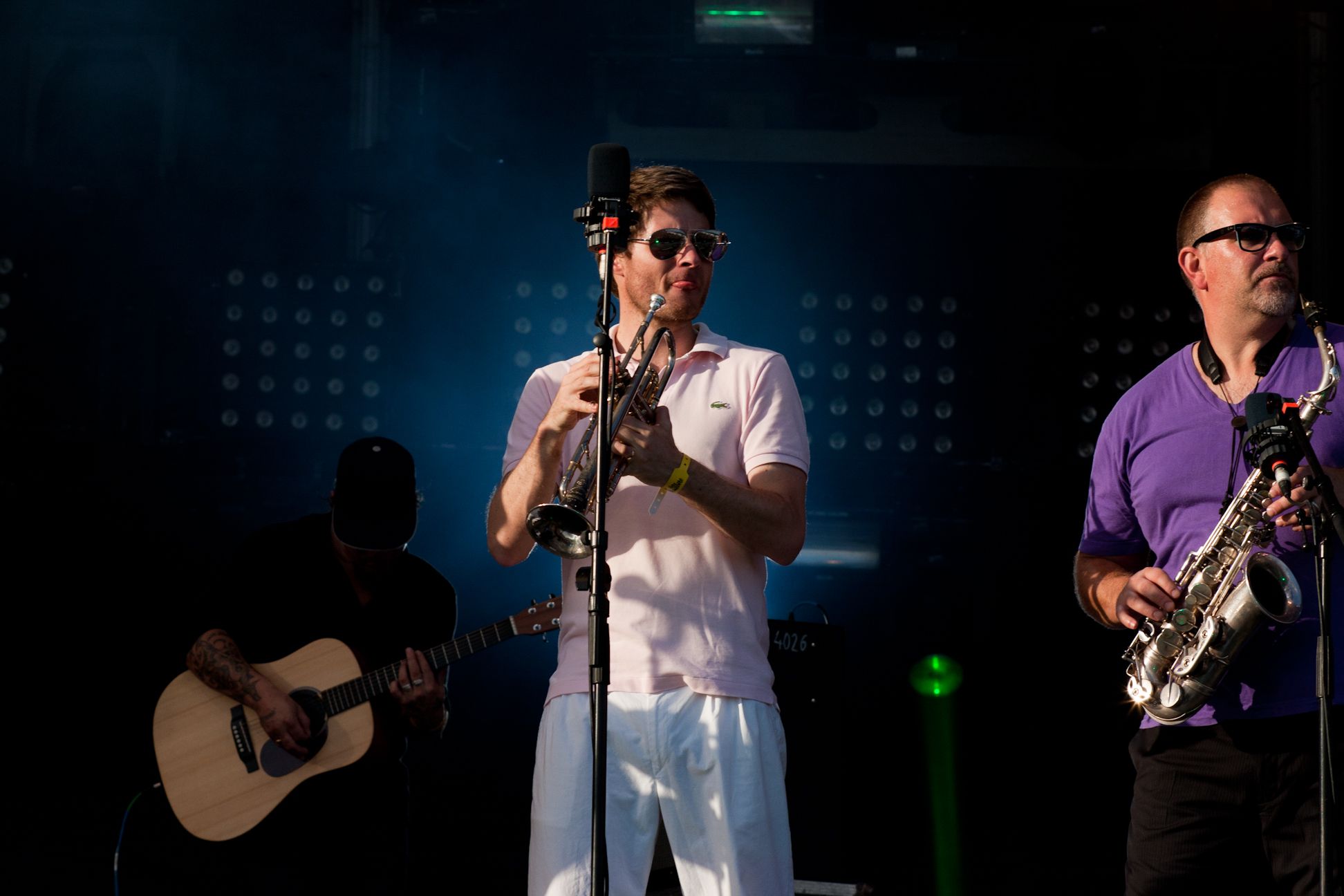 The image shows two men wearing goggles and playing saxophones on stage at a concert. There is a...