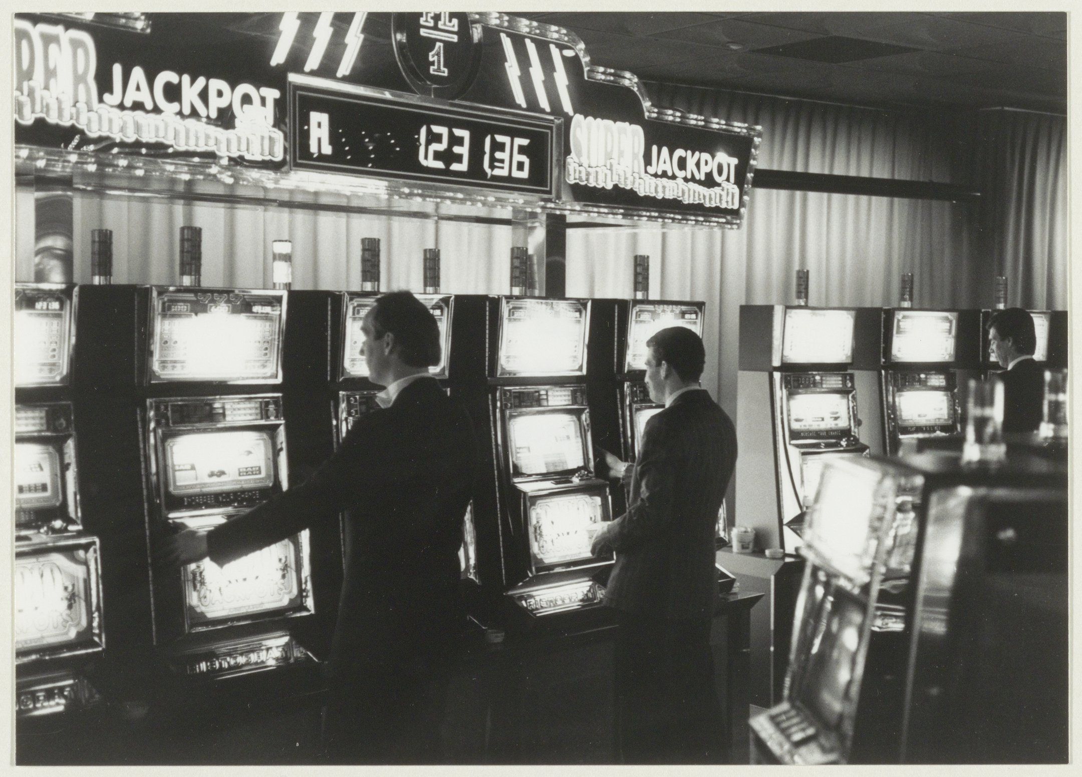 The image shows a black and white photo of two men playing slot machines in a casino. The men are...