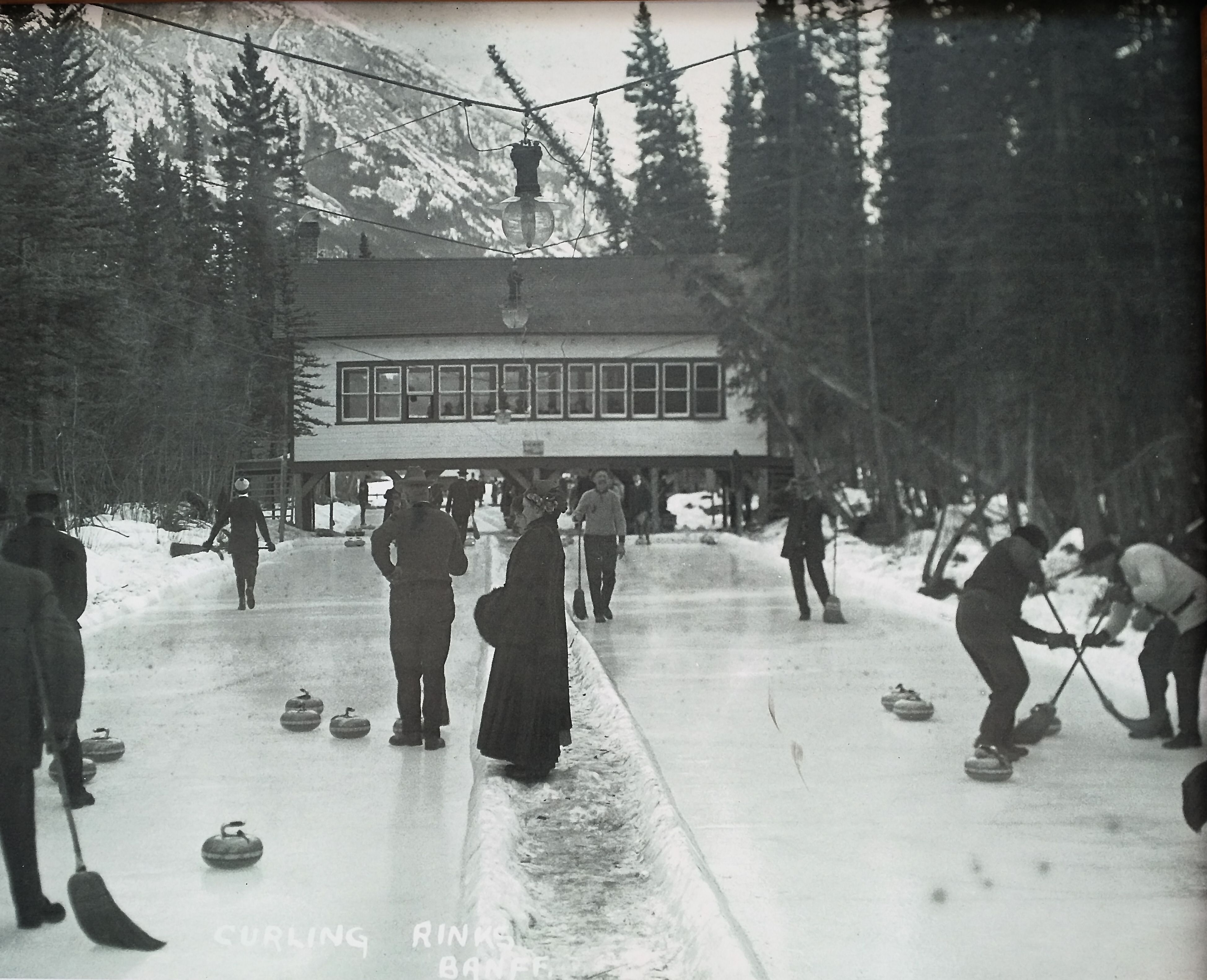 The image shows a group of people playing curling on an ice rink surrounded by trees and a building...