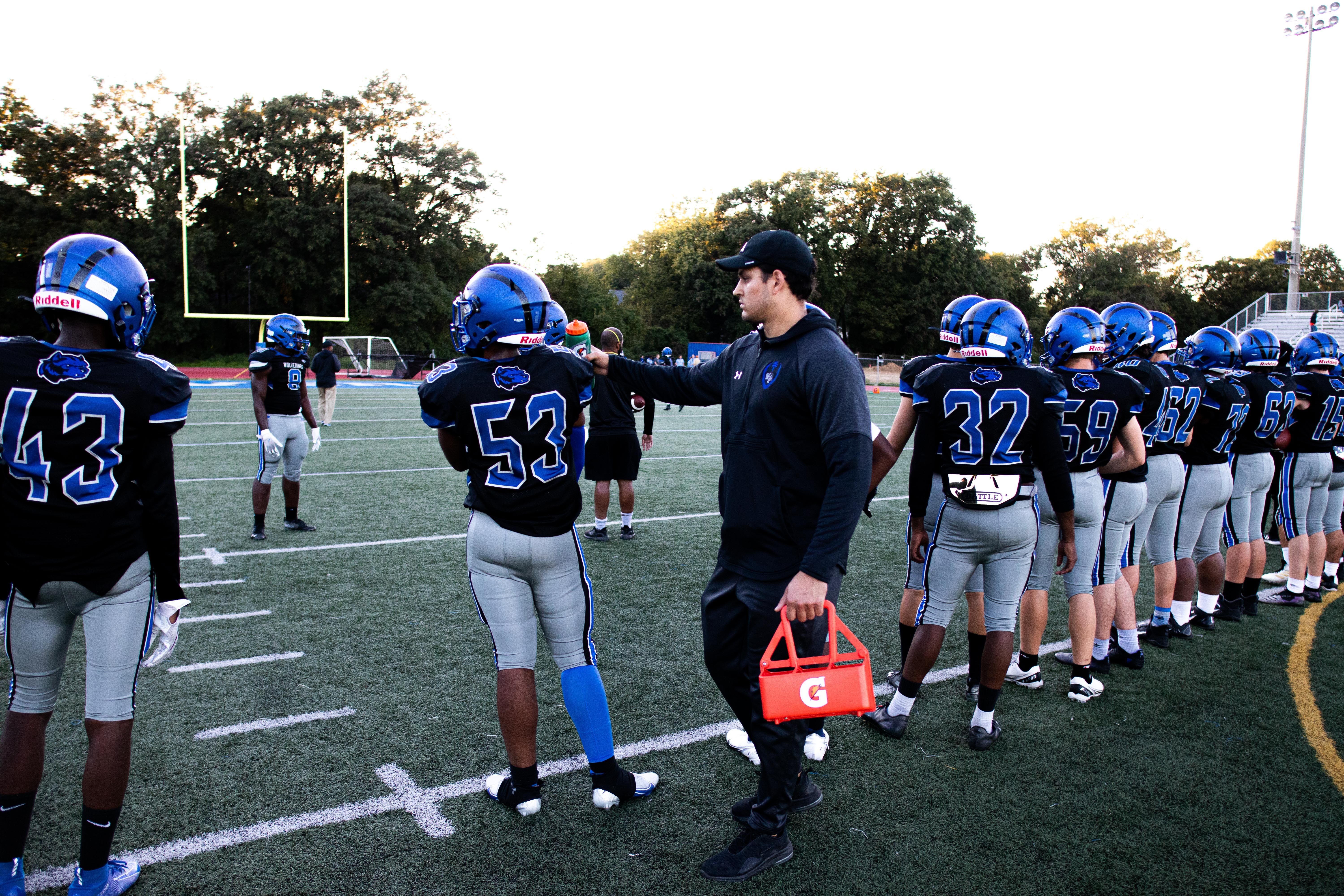 The image shows a football coach talking to his team on the field. He is wearing a cap and holding...