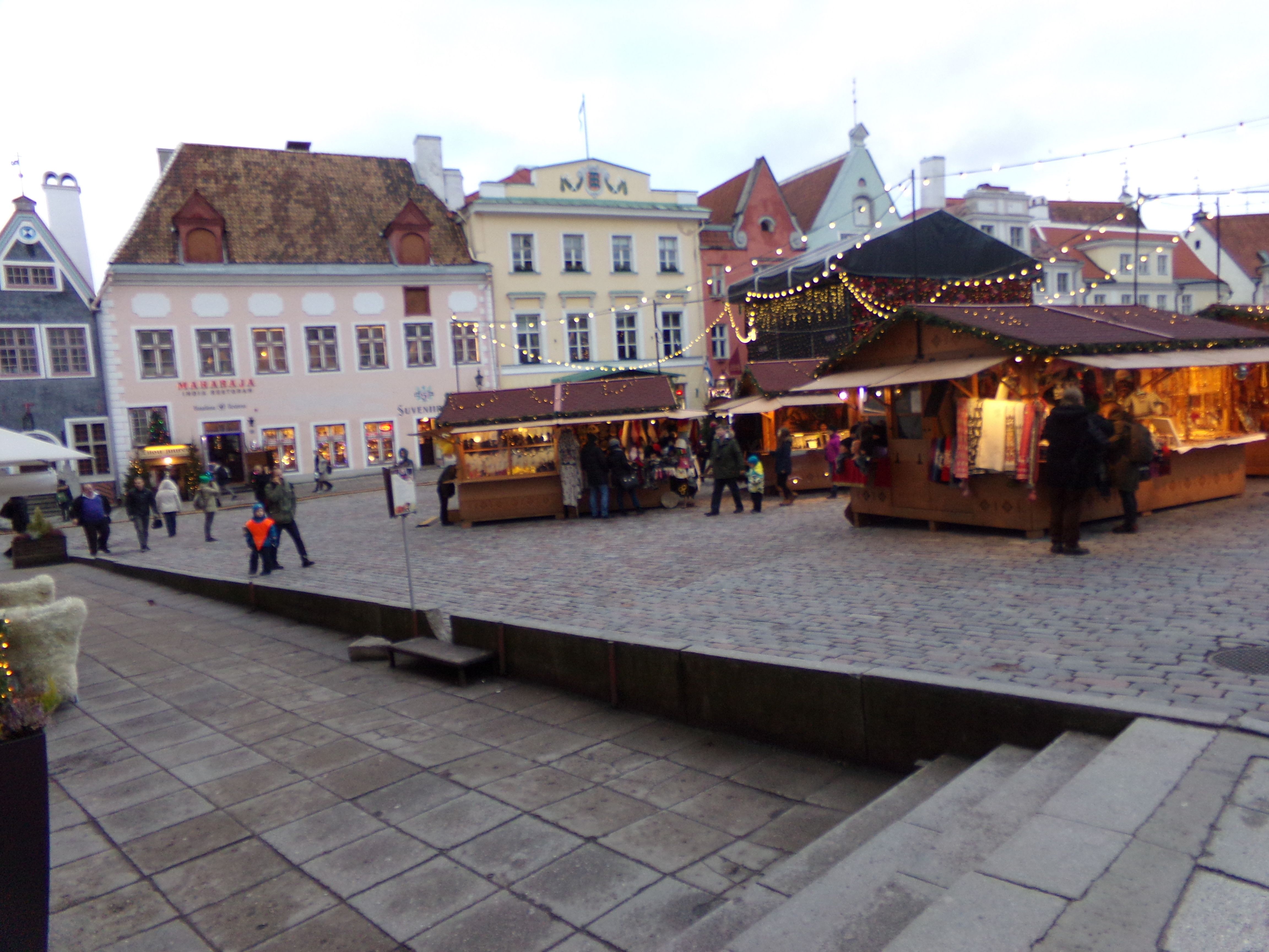 The image shows a bustling Christmas market in Tallinn, Estonia. There are many people gathered...
