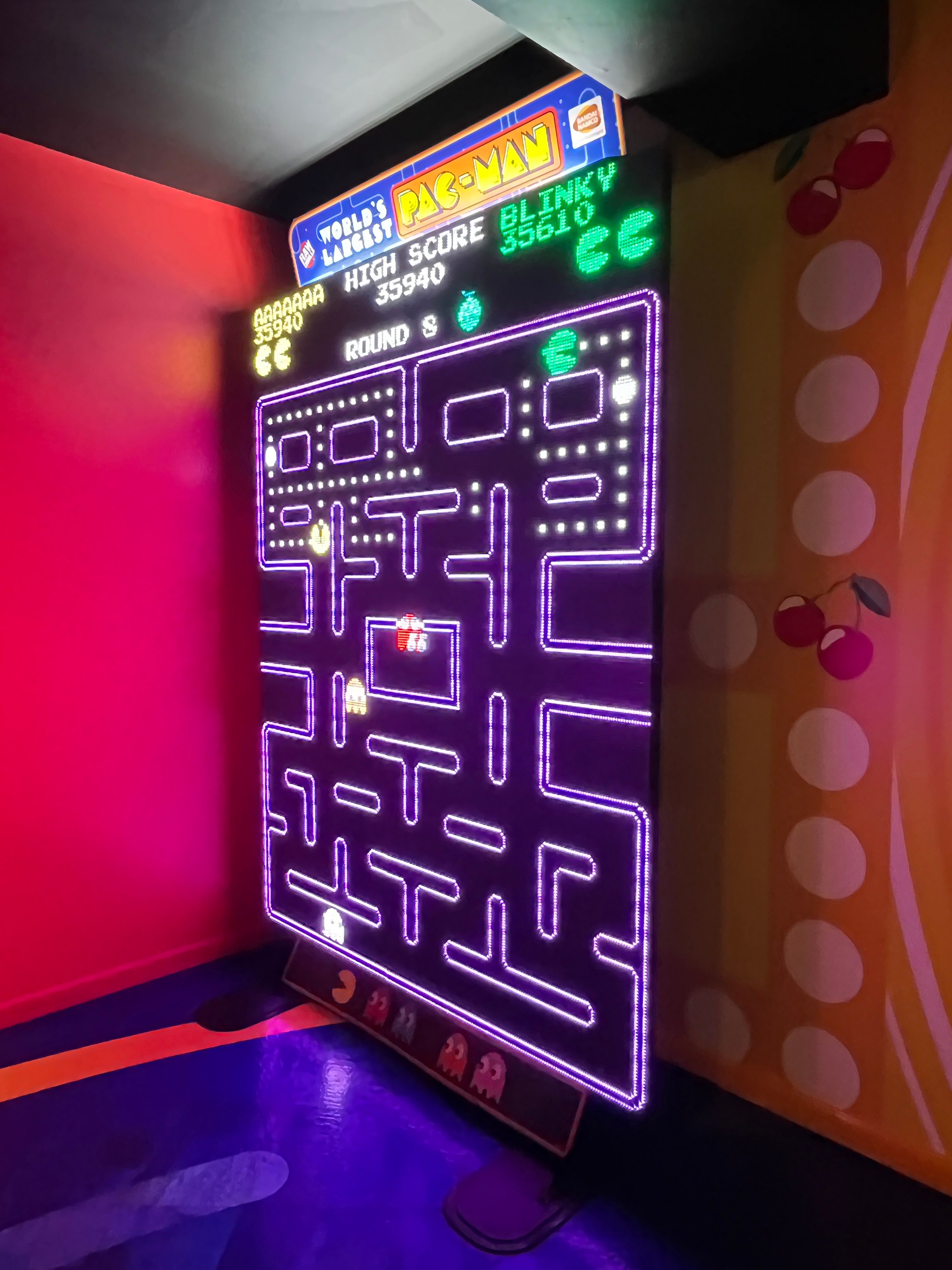 The image shows a small arcade machine with a neon sign on top of it, featuring a colorful board...
