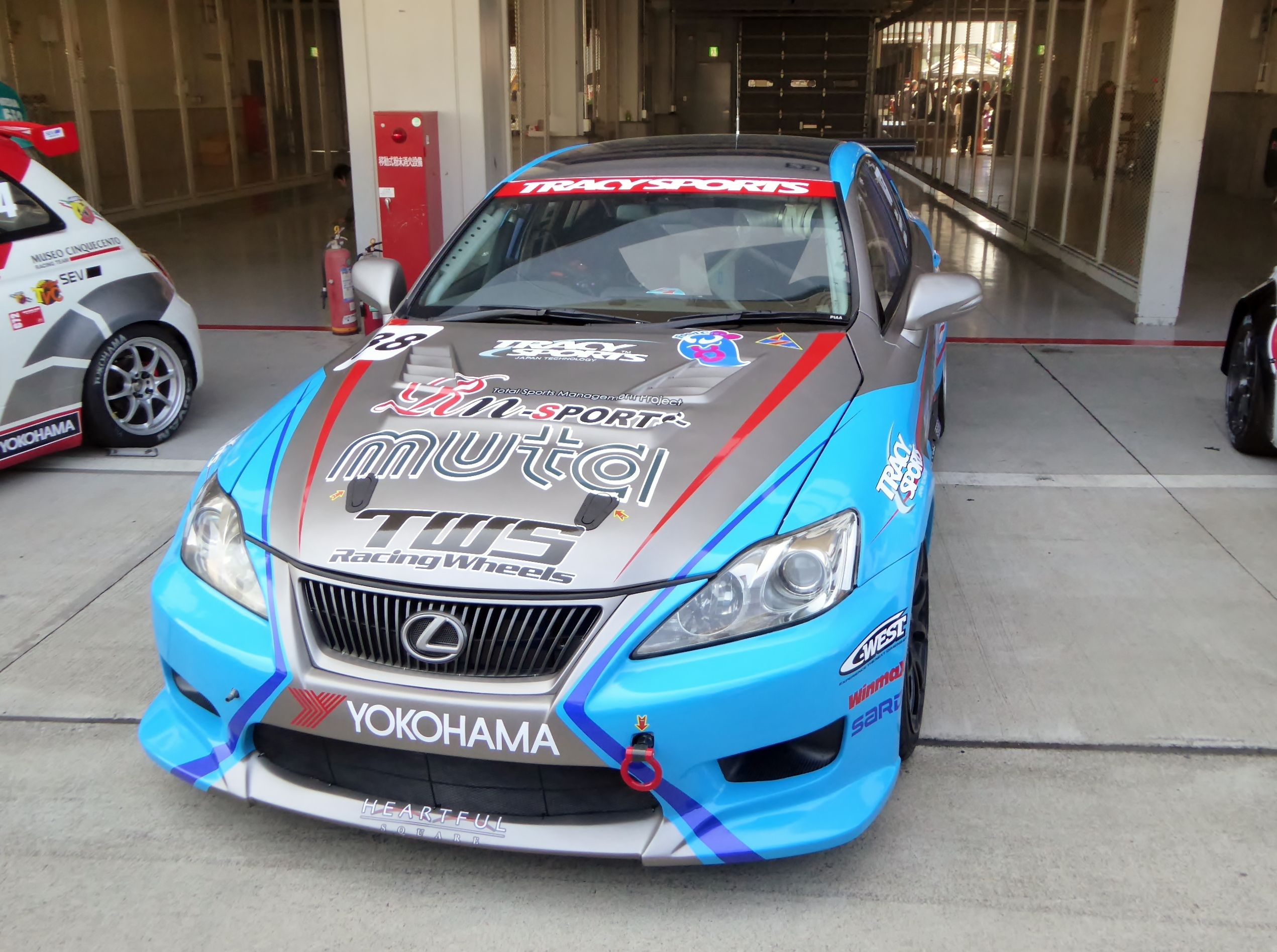 The image shows a Lexus IS F race car parked in front of a building with glass walls and pillars....