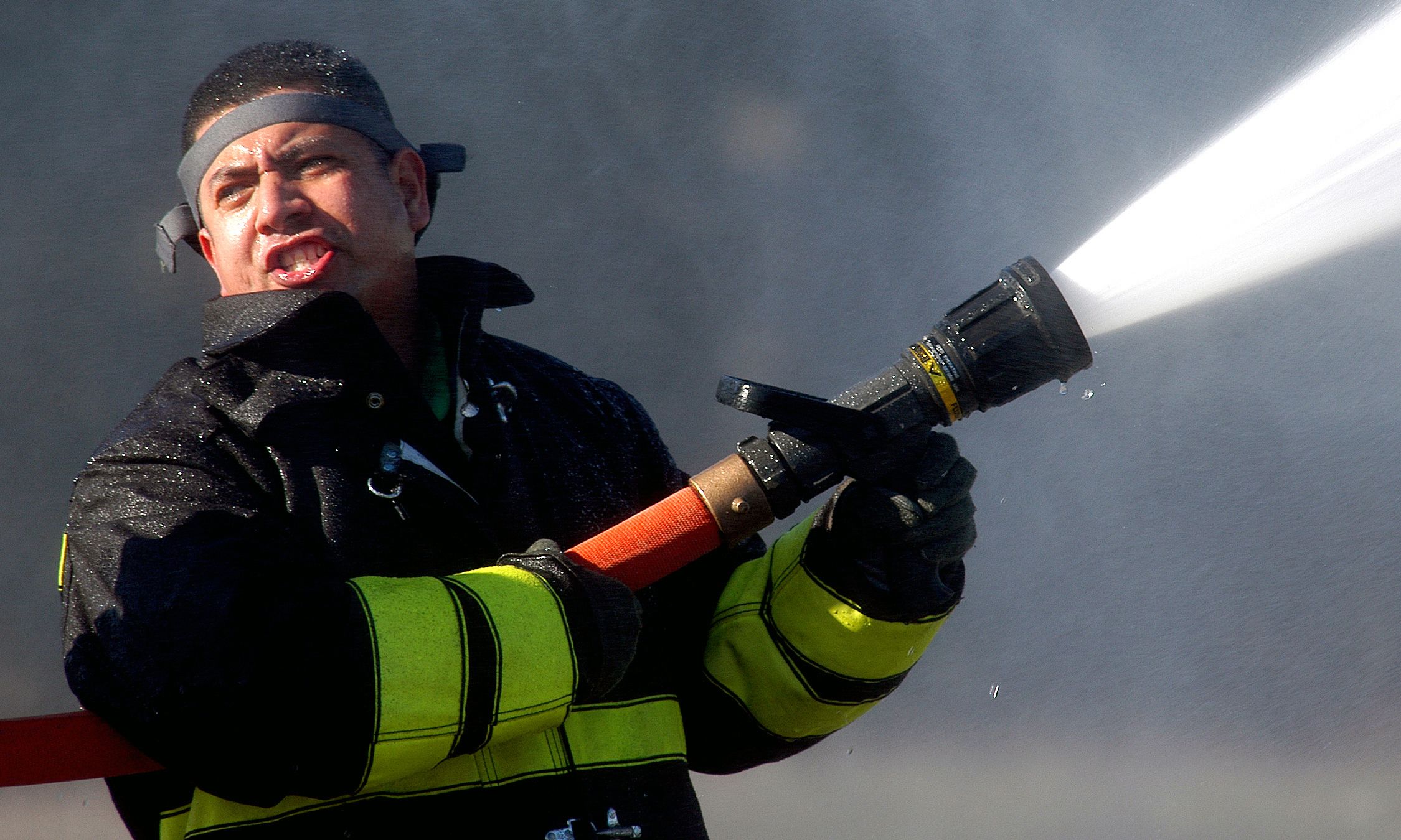 The image shows a firefighter wearing a black jacket, gloves, and a cap, holding a hose and...