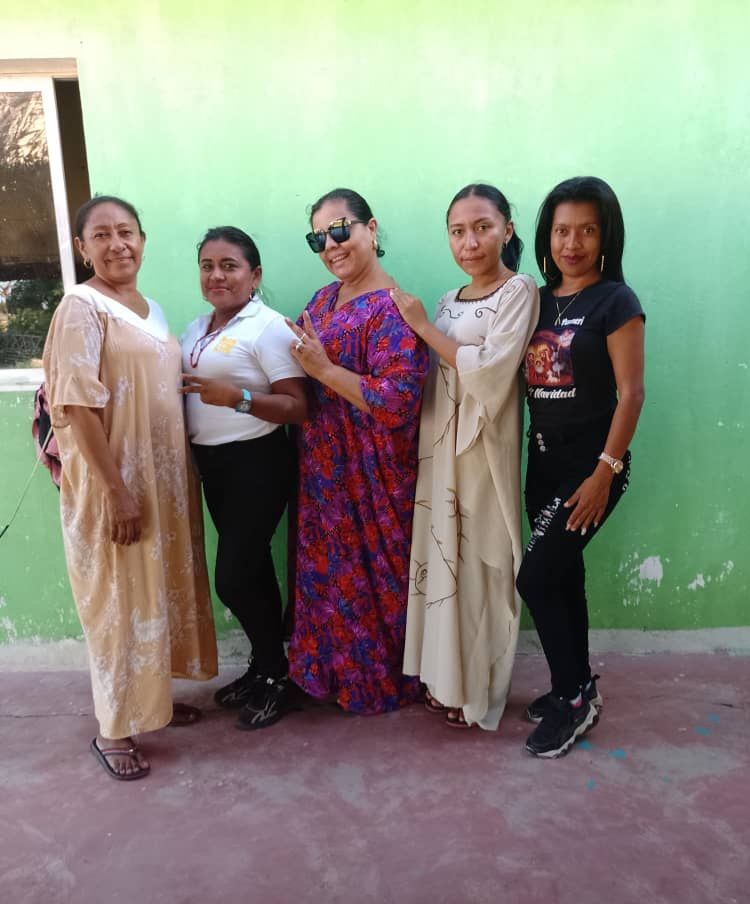 The image shows a group of women standing next to each other in front of a green wall, with a...