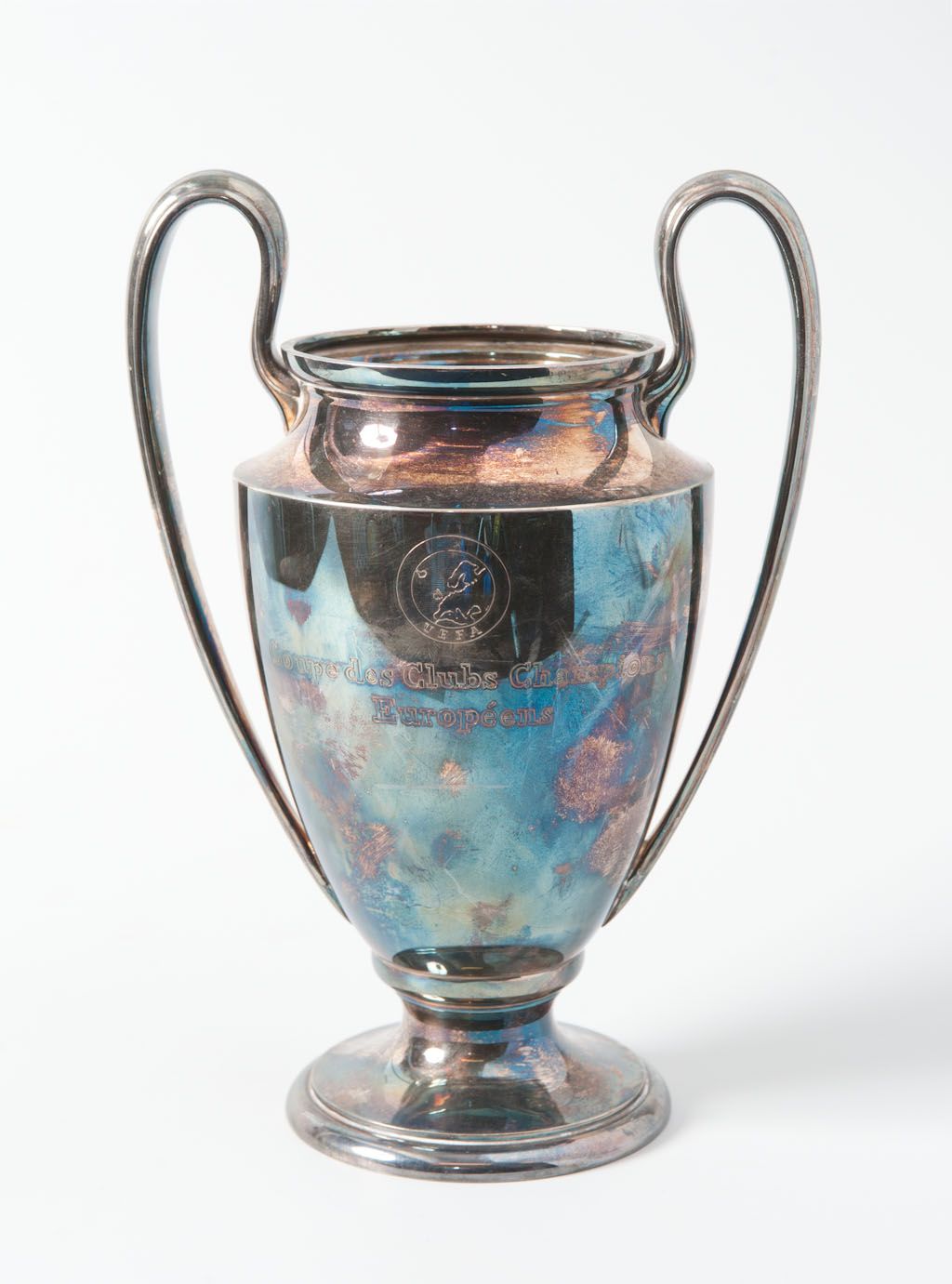 The image shows the UEFA Champions League Trophy, a steel cup with two handles and a blue and...