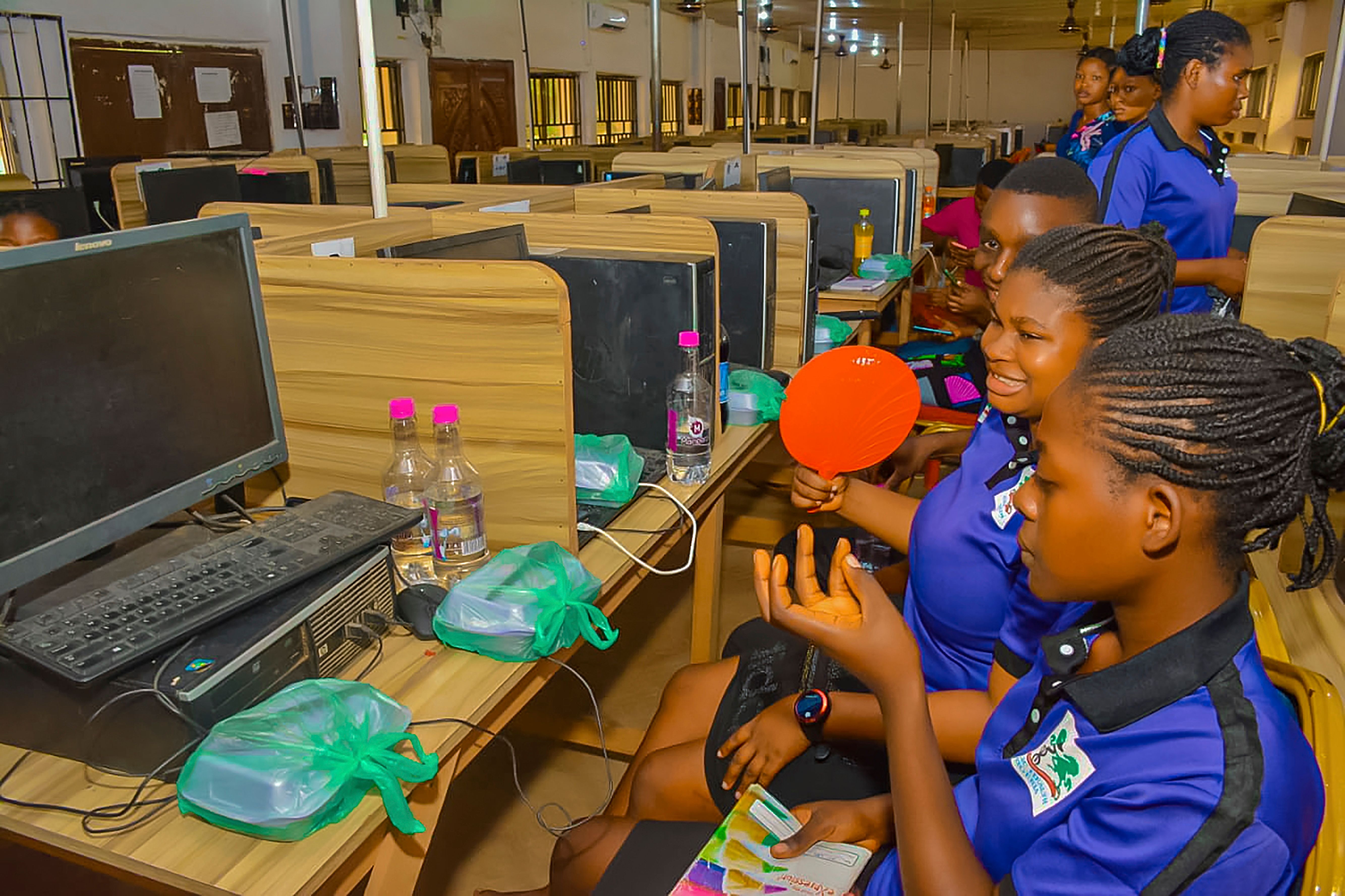 The image shows a group of young girls sitting at desks in front of computers, with monitors,...