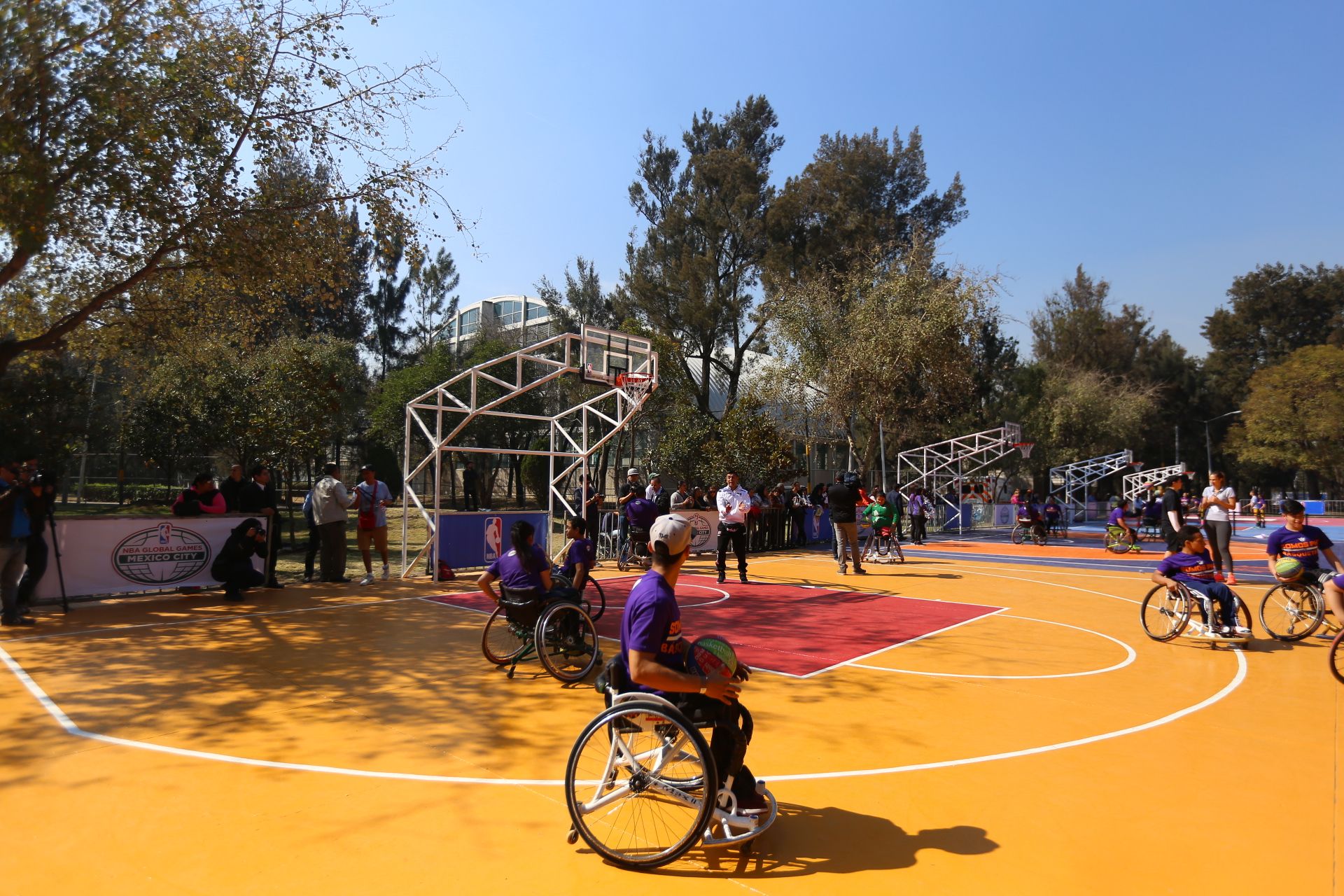The image shows a group of people in wheelchairs playing basketball on a court surrounded by trees,...