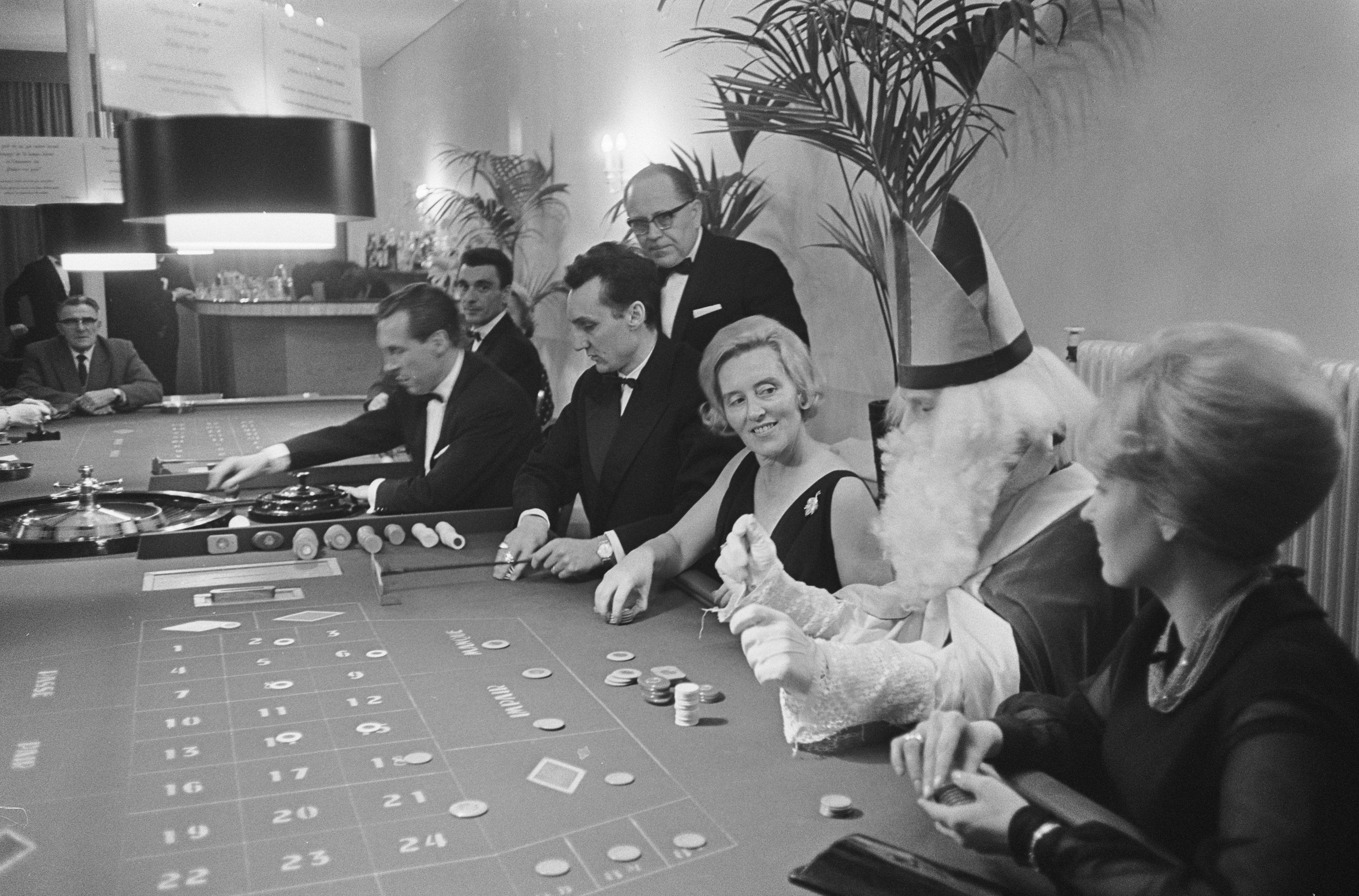 The image shows a group of people sitting around a roulette table in a casino. The table is black...