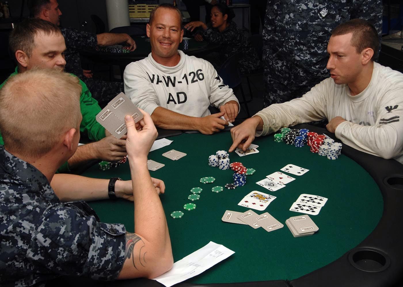 The image shows a group of men sitting around a table playing poker, with coins and cards spread...