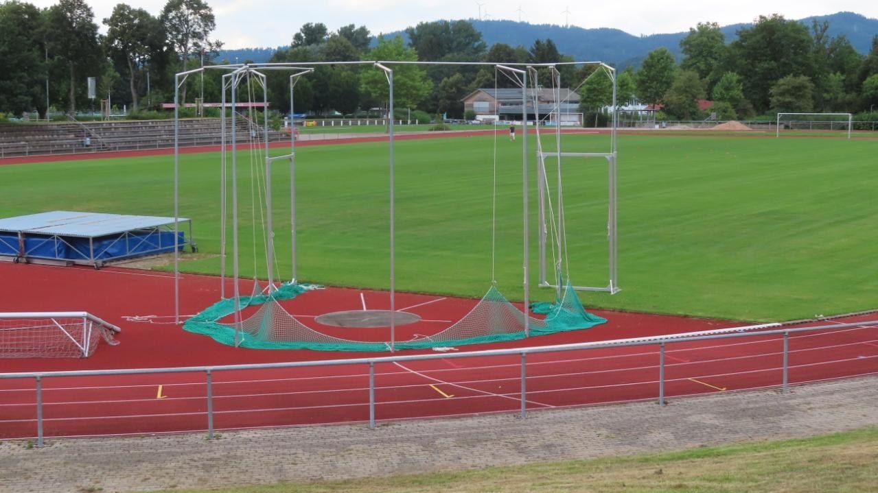 The image shows a soccer field with a net on top of it, surrounded by a metal fence, grass, a group...
