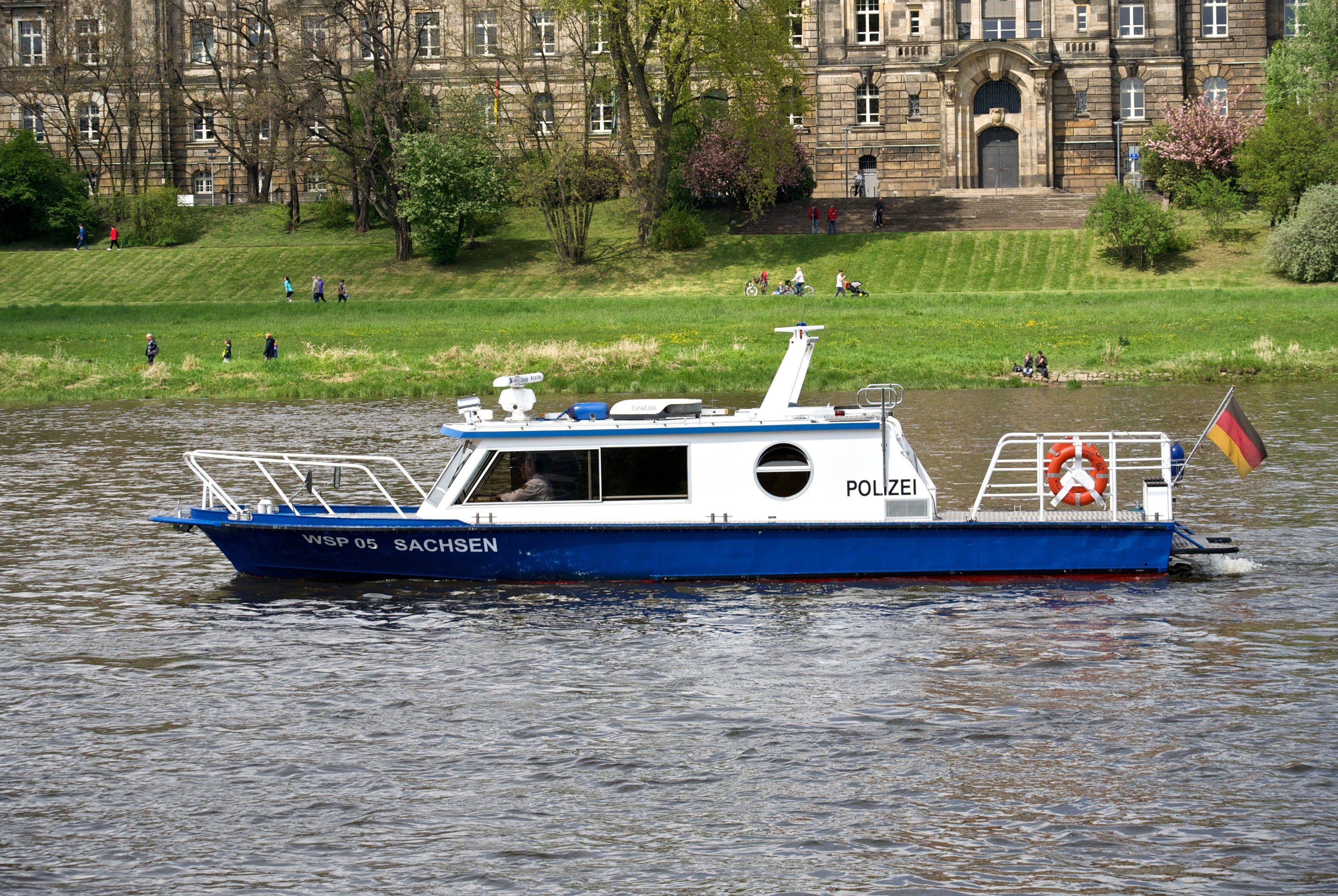 The image shows a blue and white boat traveling down a river, with a flag flying from the mast. In...