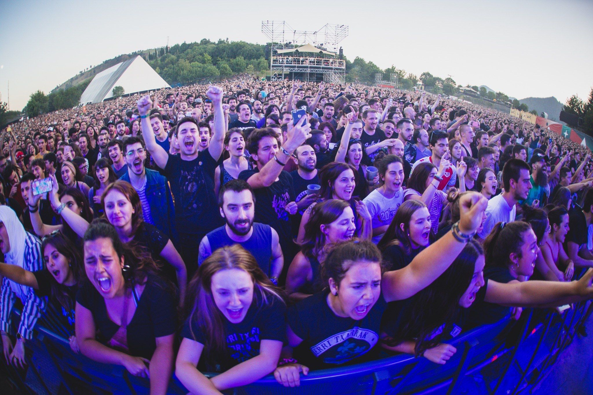 The image shows a large crowd of people at a music festival with their hands in the air, standing...