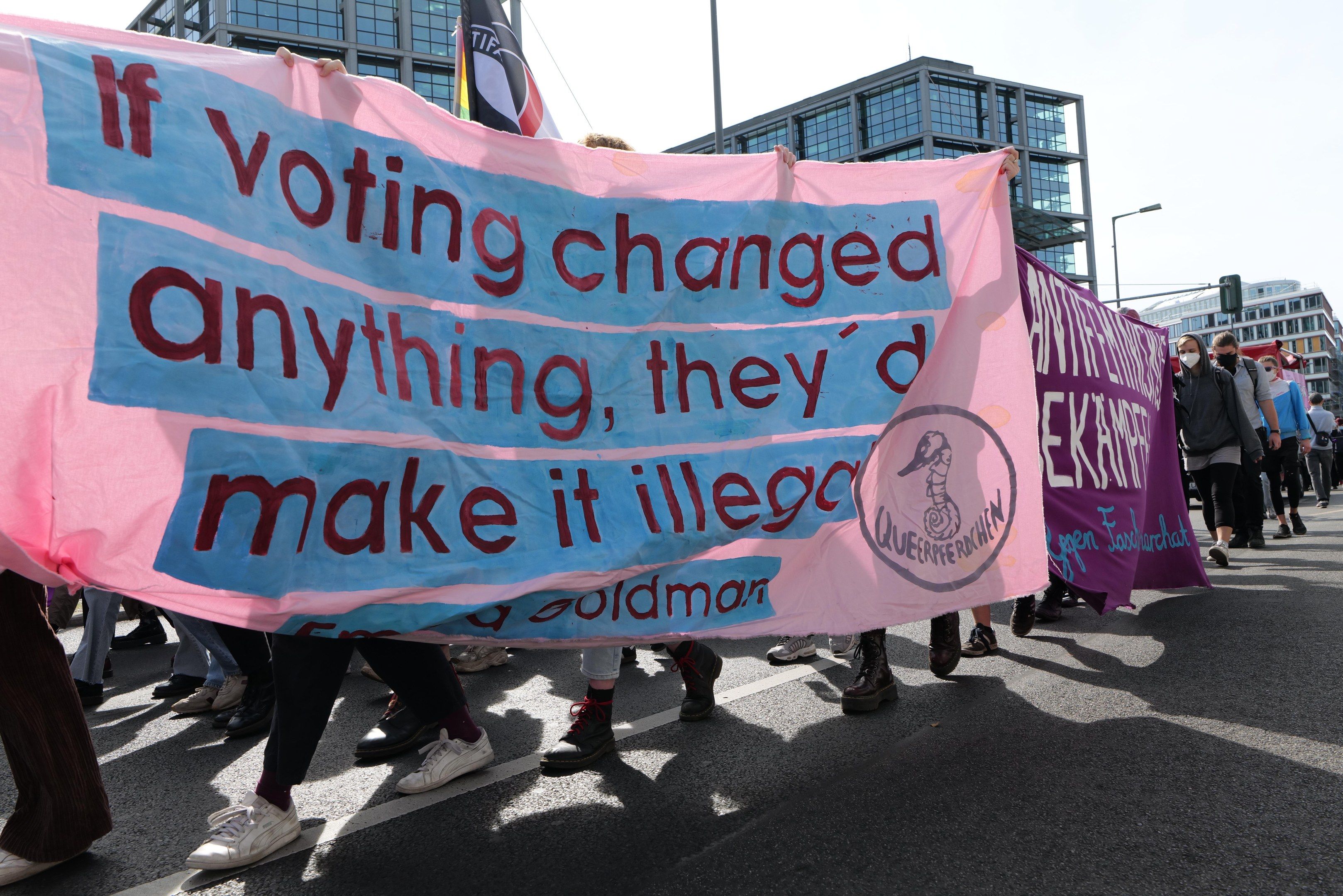 The image shows a group of people walking down a street, holding a banner that reads "If voting...