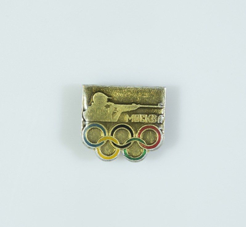 The image shows a gold and silver pin with the Olympic rings on it, set against a white background....