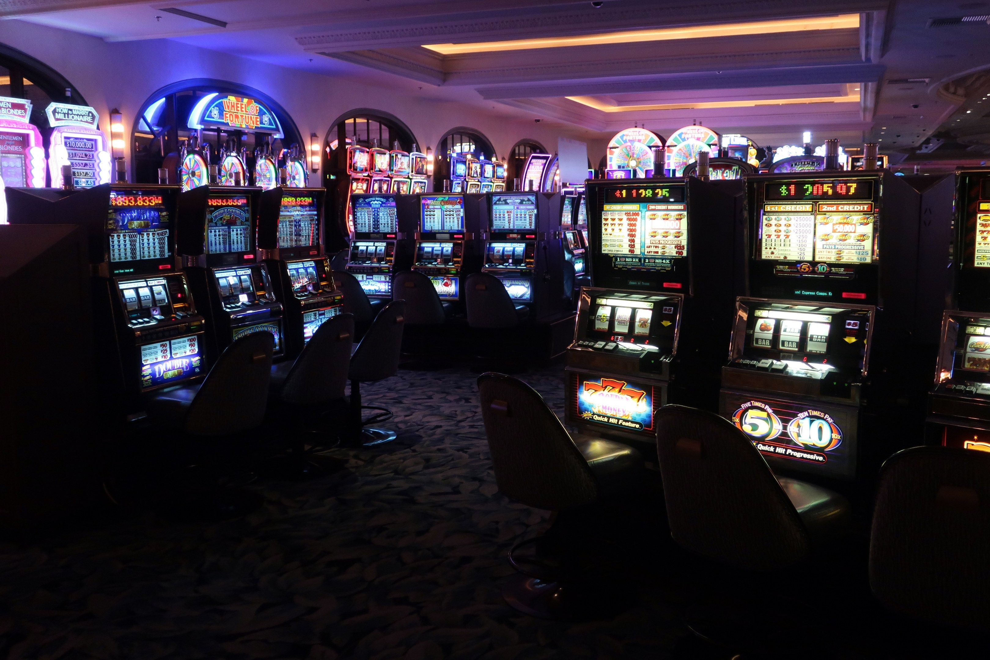 The image shows a row of slot machines in a casino, with chairs on the floor and lights on the...