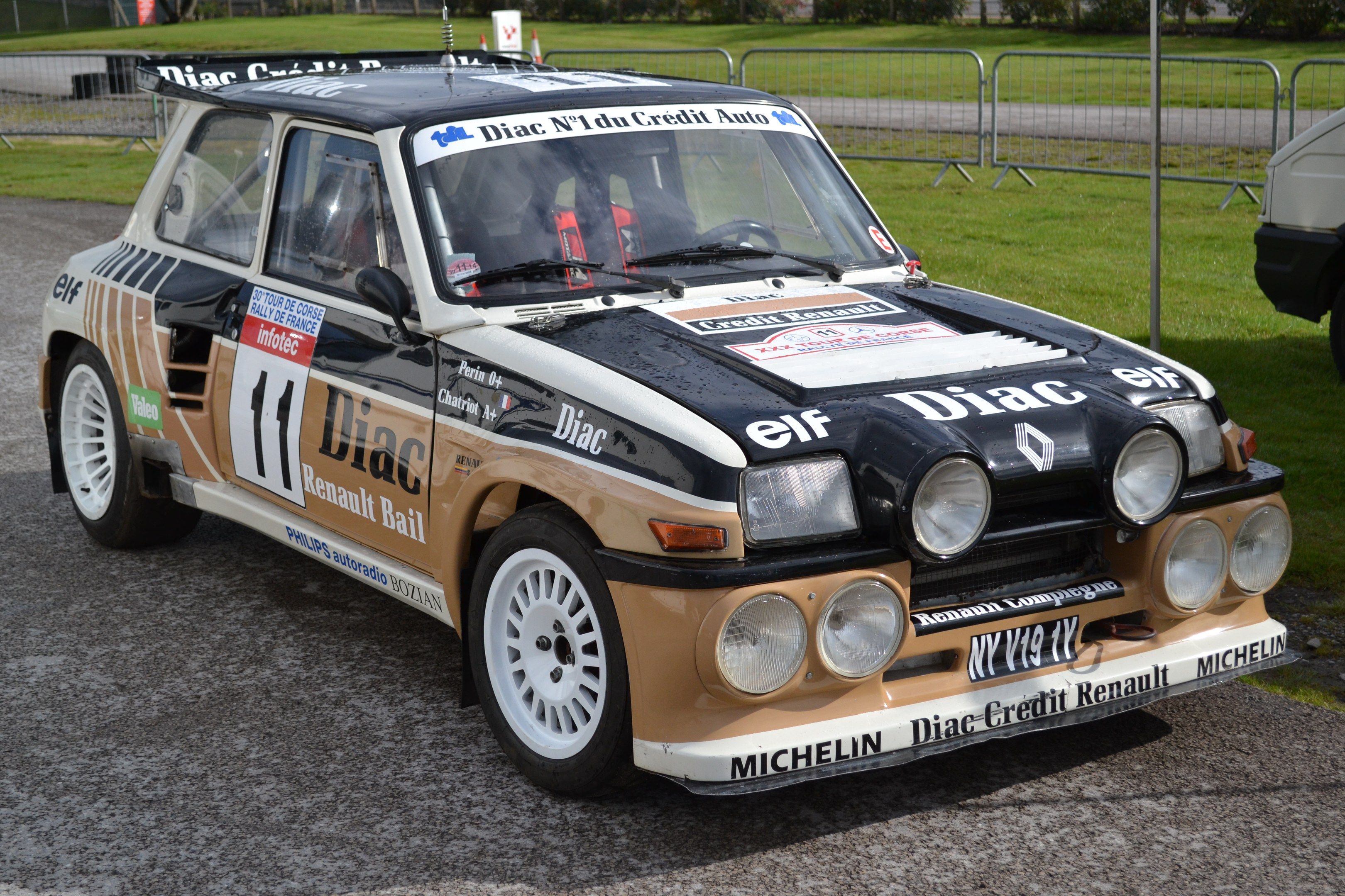 The image shows a Renault 5 Turbo RS Rally Car on the road surrounded by grass, railings, poles,...