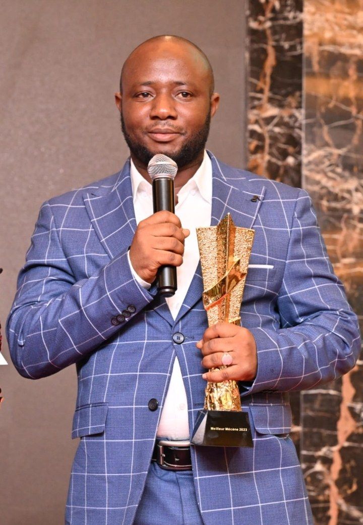 The image shows a man in a blue suit holding a microphone and a trophy, with a wall in the...