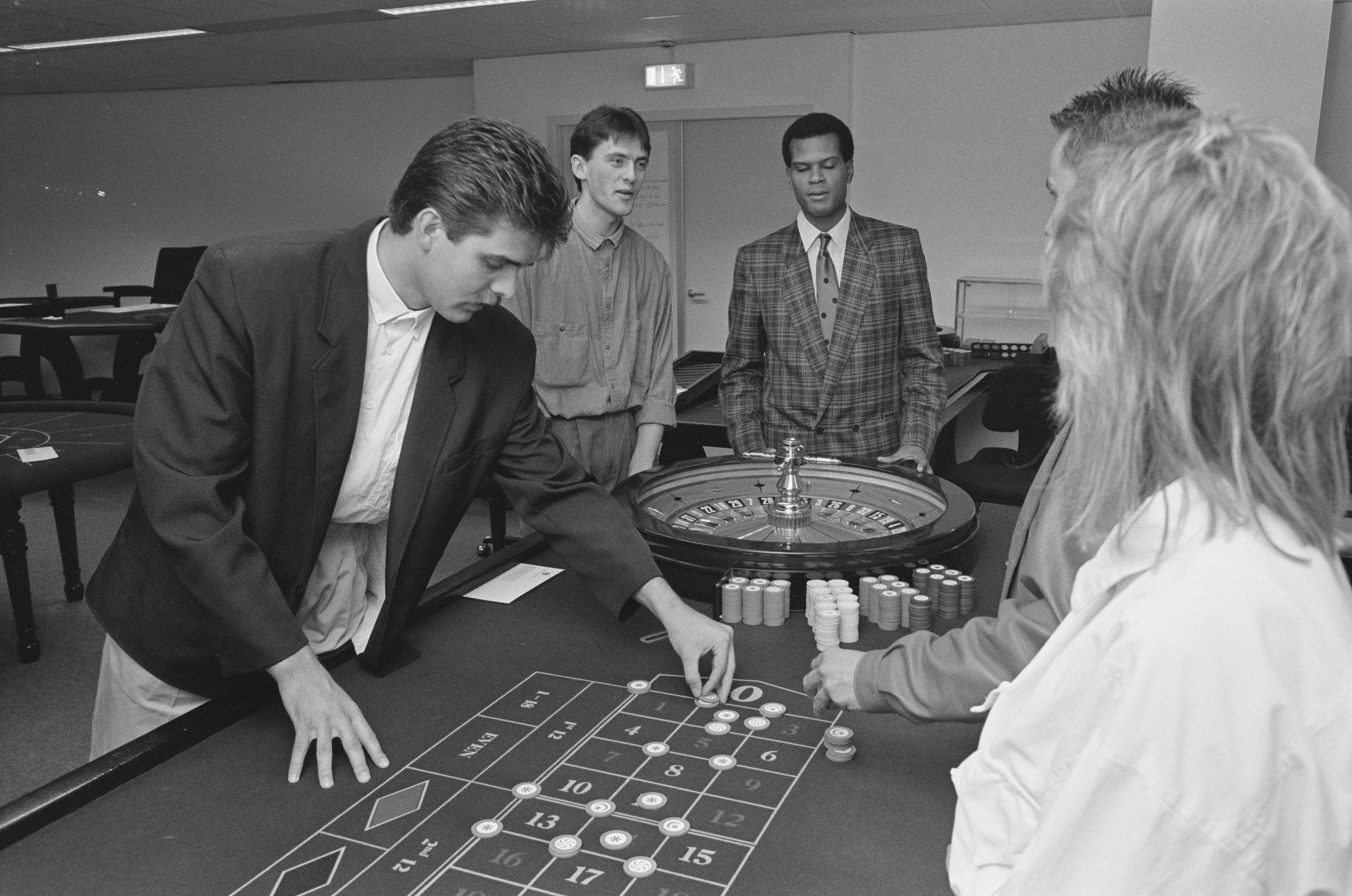 The image shows a group of people playing roulette in a casino. There are four people standing...