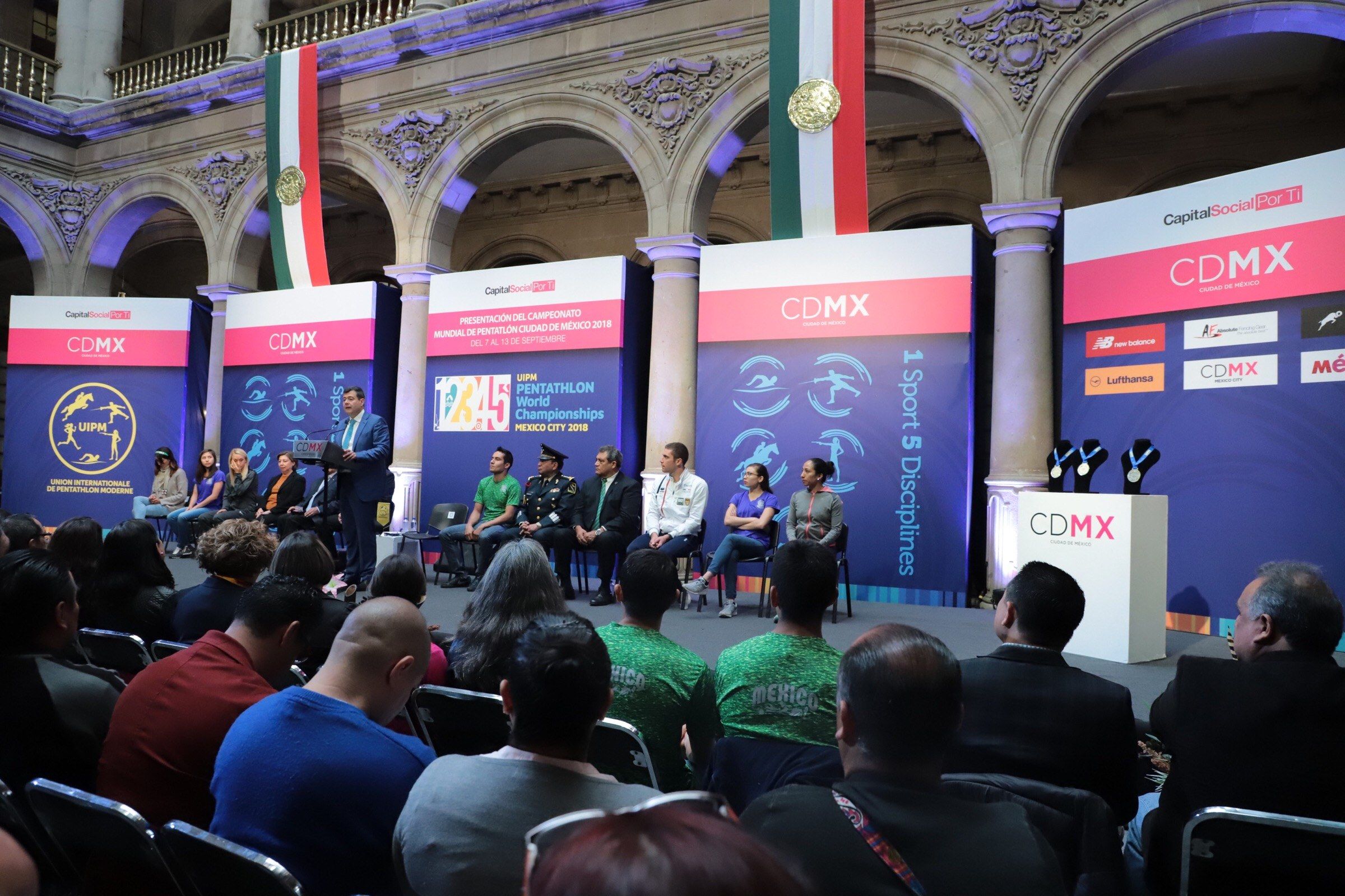 The image shows a group of people sitting on chairs in front of a stage, with a man standing beside...