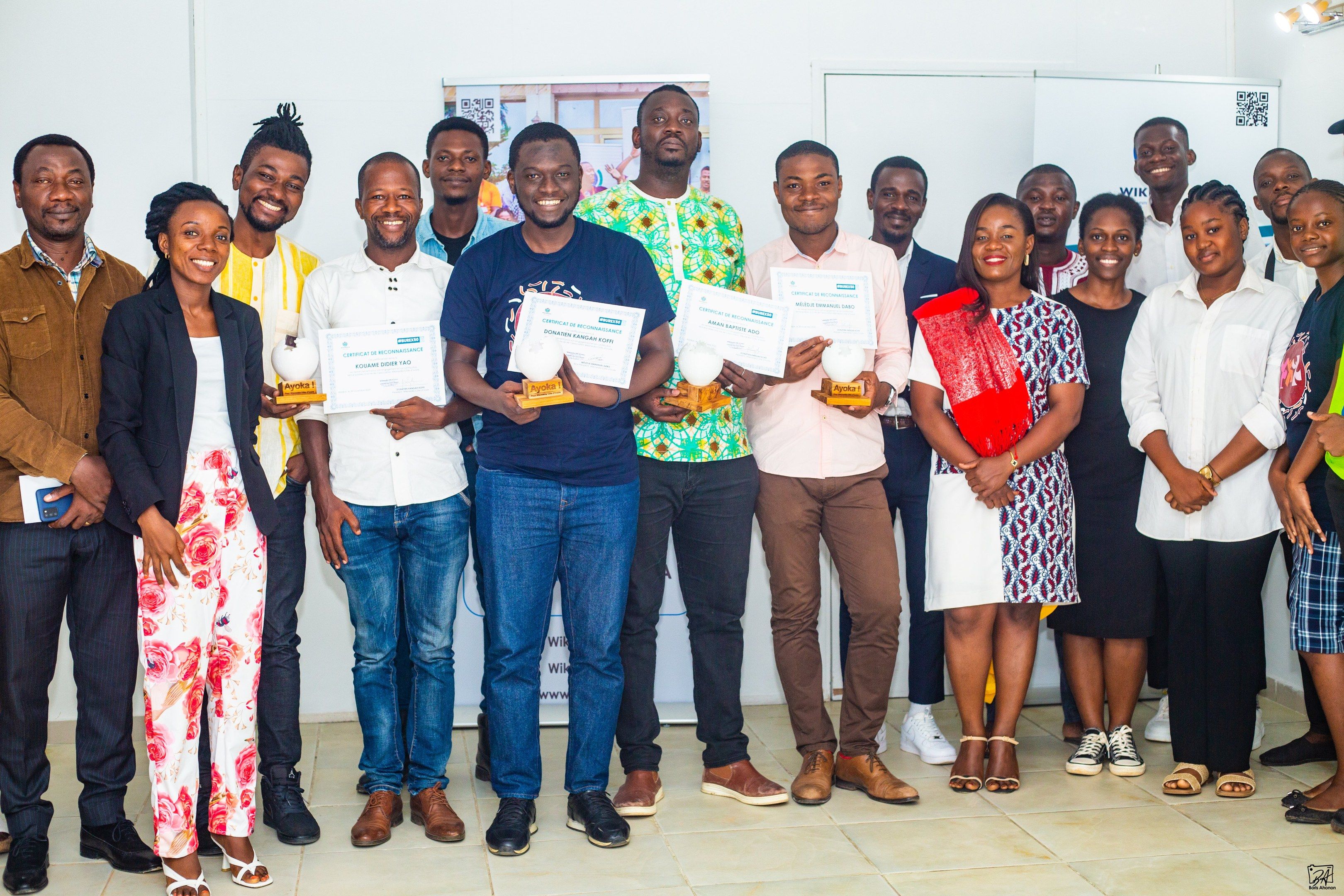 The image shows a group of people standing next to each other in a room, smiling and holding...