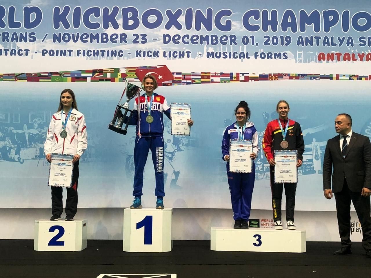 The image shows a group of five people standing on top of a podium, each holding a trophy and...