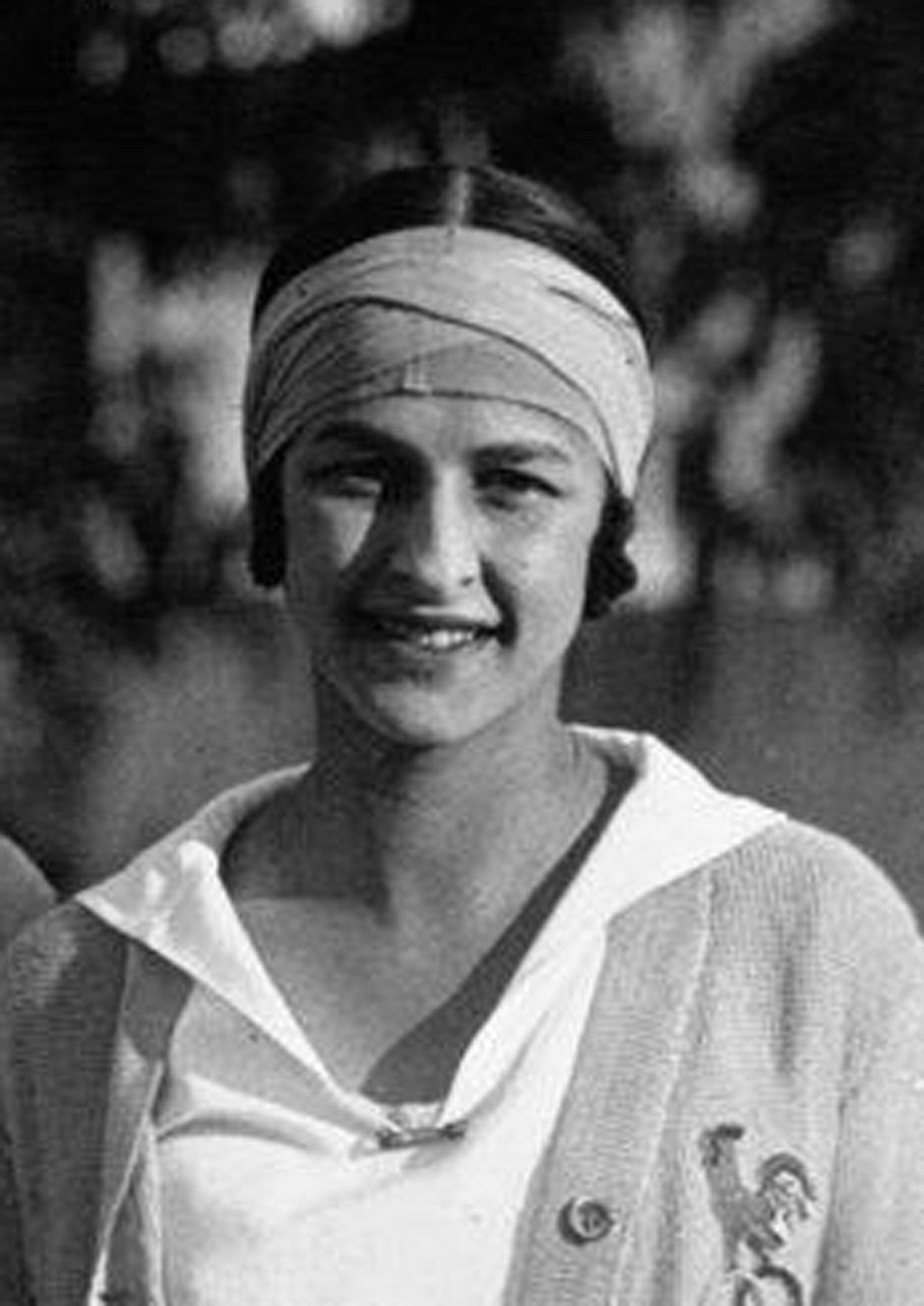 The image shows a black and white photo of a woman with a turban on her head, smiling, with a...