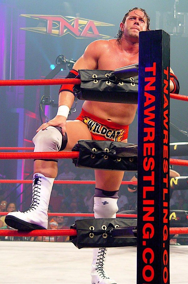 The image shows a man standing in a wrestling ring with his hands on his knees, surrounded by ropes...