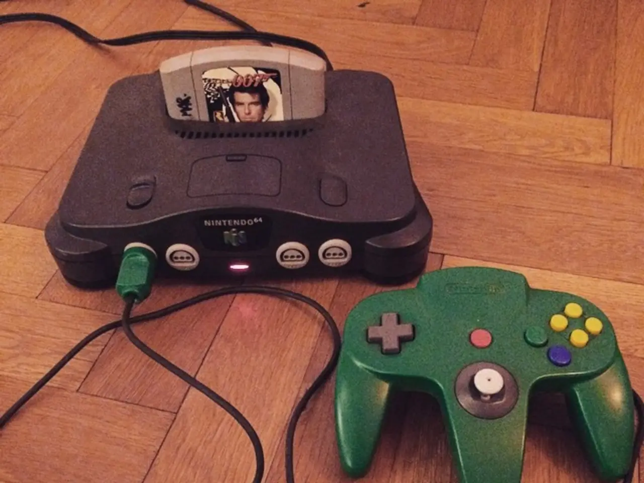 In this picture we can see a green color game controller, cables and a game cartridge on the wooden...