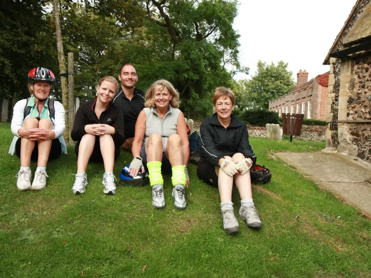 In this image we can see one man and four women are sitting on the grassy land. Behind them trees,...