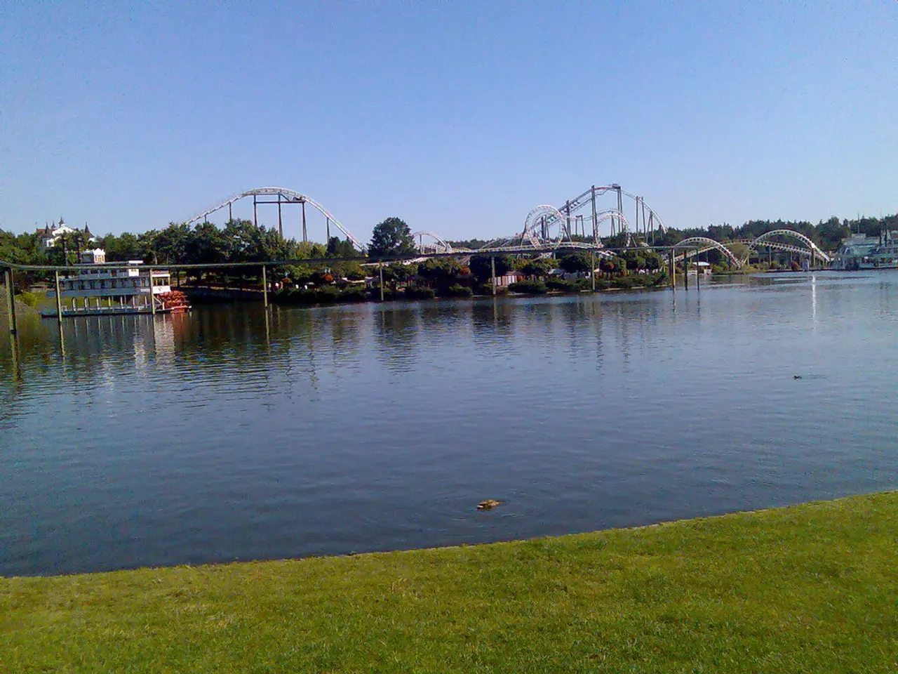 In this image we can see the roller coaster, a group of trees, buildings, some poles, a large water...