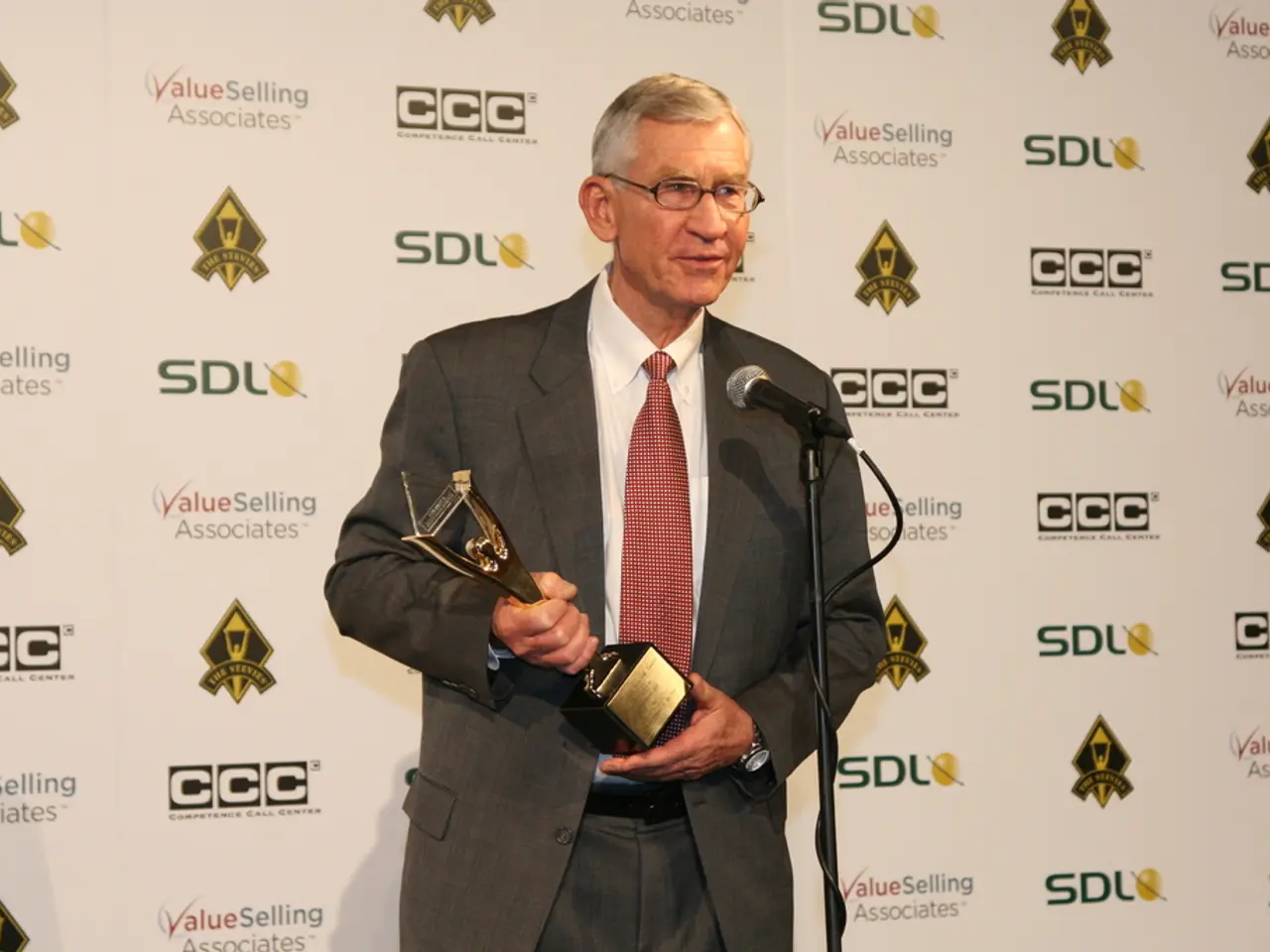 In this image we can see a man standing in front of a mike and holding a trophy with his hands. He...