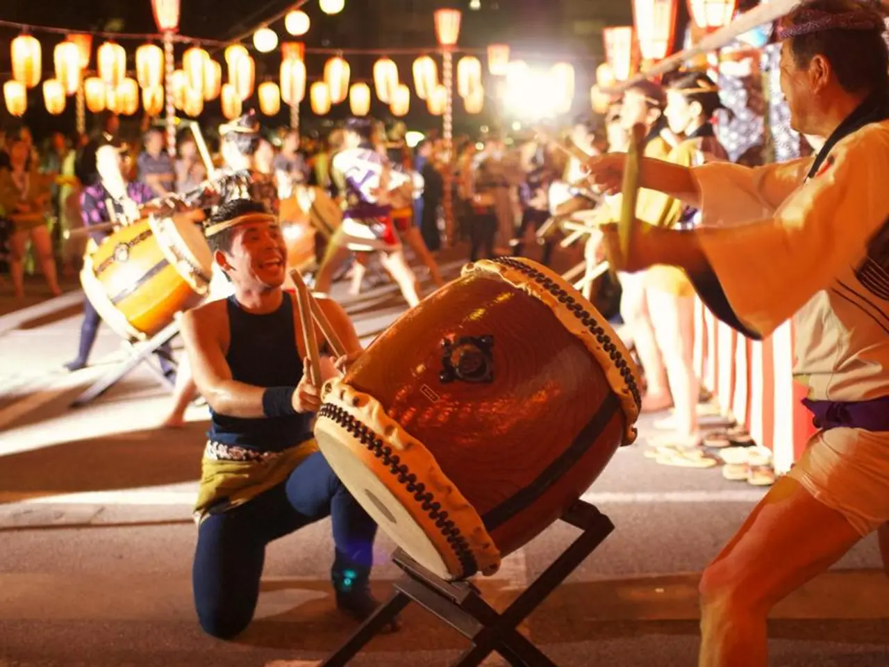 In the image there are men playing drum,it seems to be a festival and there are lights hanging on...