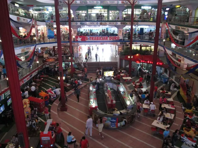 This image is taken in the mall. In this image we can see pillars, persons, tables, floors, flags,...