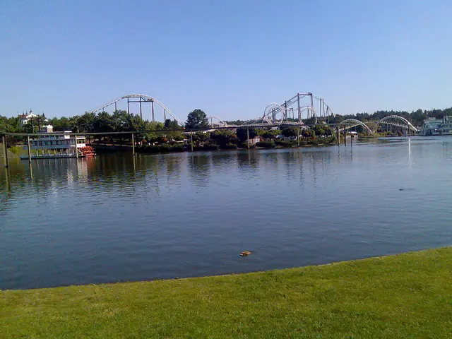In this image we can see the roller coaster, a group of trees, buildings, some poles, a large water...