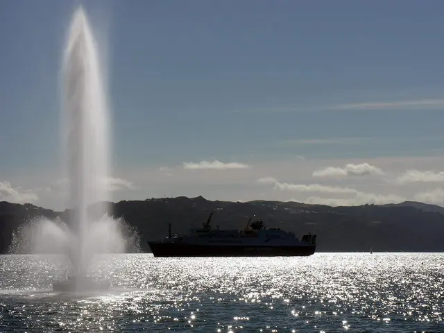 In this picture I can see a ship on the water and I can see water fountain and hills in the back...