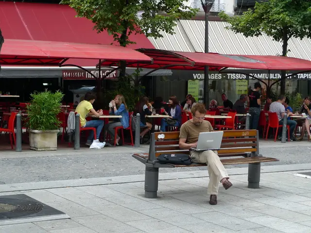 In the image there is a man sat on a bench on the street using a laptop,the picture is clicked on a...