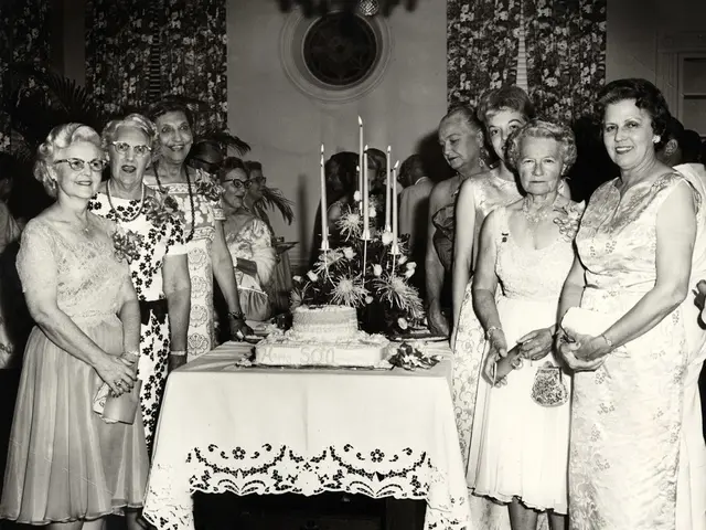 This is a black and white image. In this image we can see many women standing. There is a table. On...