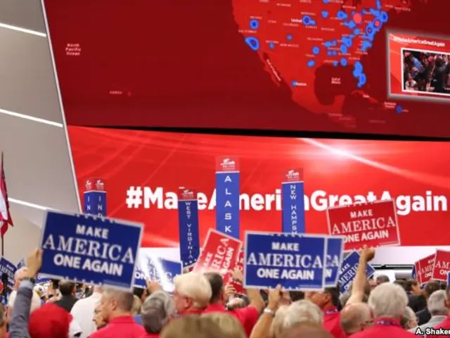The image shows a large crowd of people holding signs in front of a large screen, with flags on the...