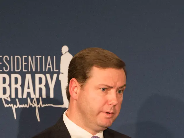 The image shows a man in a suit and tie giving a speech in front of a board with something written...