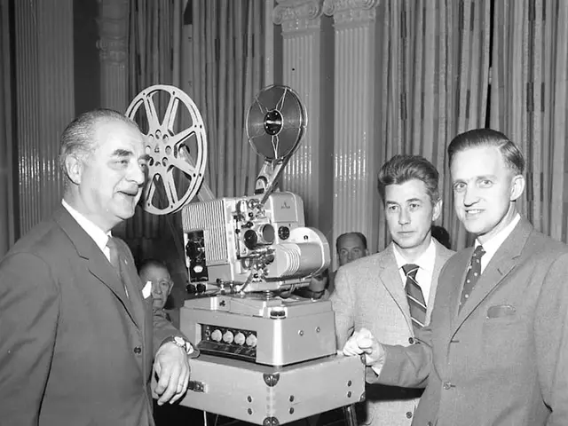 The image shows two men standing next to each other in front of an old movie projector. They are...