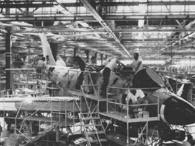 The image shows a black and white photo of an airplane being built in a factory. There are several...