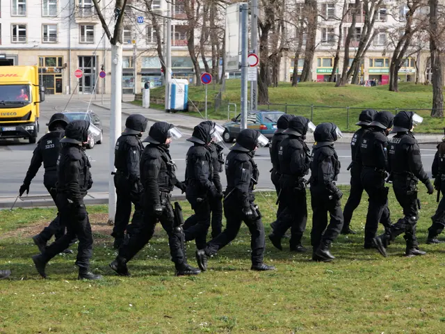 The image shows a group of police officers in riot gear walking across a grass covered field. They...