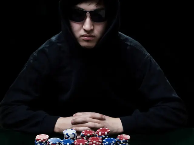 The image shows a man wearing a black hoodie and sunglasses, sitting at a table with poker chips in...