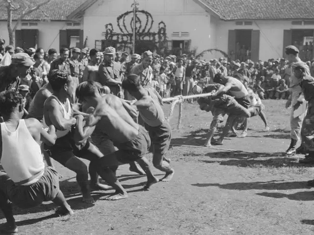 The image shows a group of men playing a game of tug of war in front of a crowd of people, some of...
