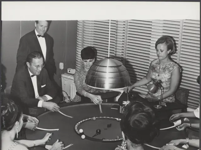 The image shows a group of people sitting around a table playing poker. There are men and women of...